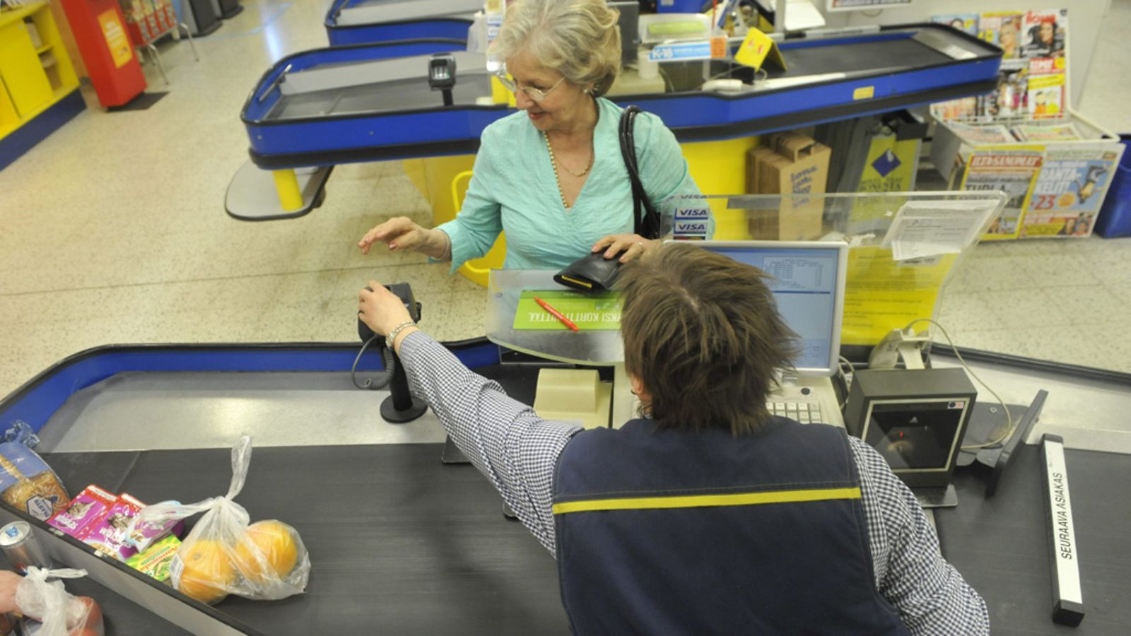 Tallenna  Myyjä opastaa iäkästä naista korttimaksussa supermarketissa.