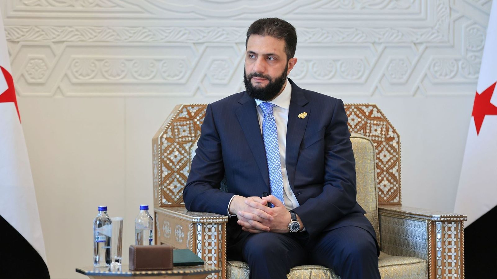 Ahmed Al-Sharaa istuu puku päällään koristeellisessa tuolissa, Syyrian lippu esillä molemmin puolin.